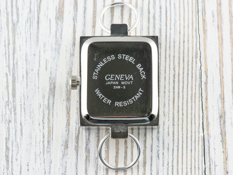 K&ouml;nnte beinhalten: Silberne quadratische Uhr mit einem Edelstahlboden. Das Zifferblatt der Uhr zeigt "Geneva Japan Movt SNW-5 Water Resistant".