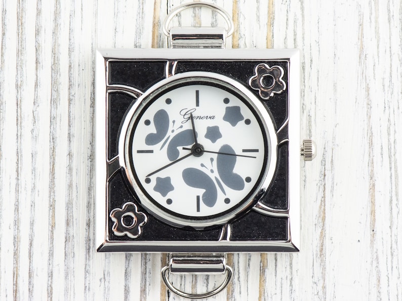May include: A silver square watch with a black face and white butterfly design. The watch face says "Geneva" and has black hands.