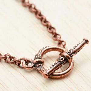 Pu&ograve; includere: Primo piano di una chiusura e catena di collana color rame. La chiusura a toggle presenta un design ornato e la catena &egrave; composta da piccole maglie rotonde. Il gioiello &egrave; posto su una superficie di legno chiaro.