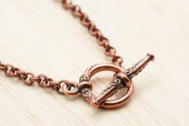 Pu&ograve; includere: Primo piano di una chiusura e catena di collana color rame. La chiusura a toggle presenta un design ornato e la catena &egrave; composta da piccole maglie rotonde. Il gioiello &egrave; posto su una superficie di legno chiaro.
