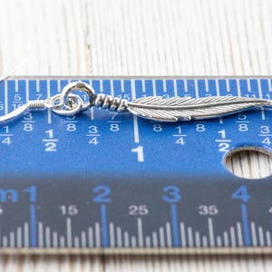Op de afbeelding: Zilveren veren oorbellen met een haak sluiting. De oorbellen worden gemeten met een liniaal, wat aangeeft dat ze ongeveer 2,5 cm lang zijn.