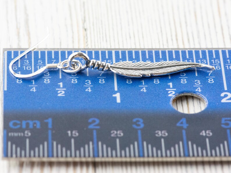 Op de afbeelding: Zilveren veren oorbellen met een haak sluiting. De oorbellen worden gemeten met een liniaal, wat aangeeft dat ze ongeveer 2,5 cm lang zijn.