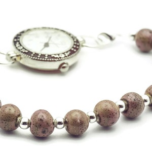 May include: A silver watch with a white face and black markings is attached to a bracelet made of brown beads with silver accents.