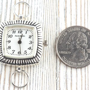 May include: A silver pocket watch with a white face and black numerals, next to a U.S. quarter for size comparison. The watch has a square face with a decorative border and the brand name "NARMI".