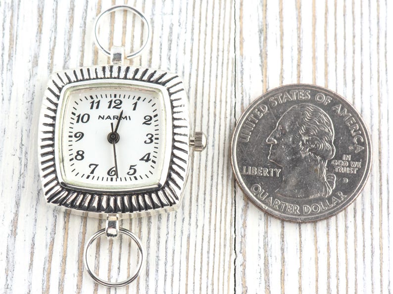 May include: A silver pocket watch with a white face and black numerals, next to a U.S. quarter for size comparison. The watch has a square face with a decorative border and the brand name "NARMI".