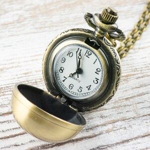 May include: A gold-toned pocket watch with a chain, open to reveal a white face with black hands and black Roman numeral markings. The watch face reads "QUARTZ".