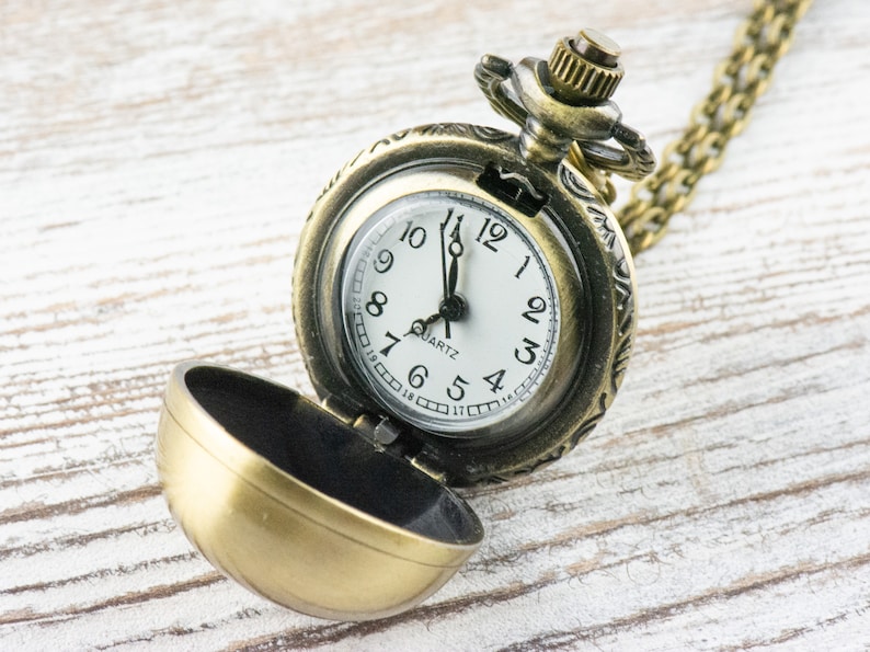May include: A gold-toned pocket watch with a chain, open to reveal a white face with black hands and black Roman numeral markings. The watch face reads "QUARTZ".
