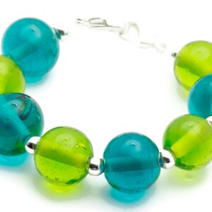 Puede incluir: Un brazalete hecho de cuentas de vidrio alternas de color verde azulado y verde. Las cuentas son redondas y tienen una superficie ligeramente texturizada. El brazalete se asegura con un cierre plateado.