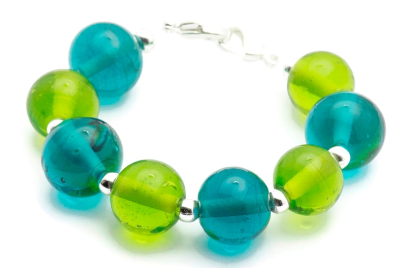 Puede incluir: Un brazalete hecho de cuentas de vidrio alternas de color verde azulado y verde. Las cuentas son redondas y tienen una superficie ligeramente texturizada. El brazalete se asegura con un cierre plateado.