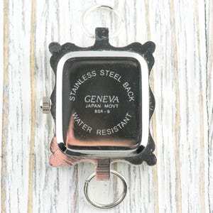 K&ouml;nnte beinhalten: Eine silberfarbene Geneva-Uhr mit rechteckigem Zifferblatt und Edelstahlr&uuml;ckseite. Die Uhr hat einen dekorativen Rahmen und einen Schl&uuml;sselring-Aufsatz. Der Text auf dem Zifferblatt lautet "Geneva Japan Movt BSR-9 Water Resistant."