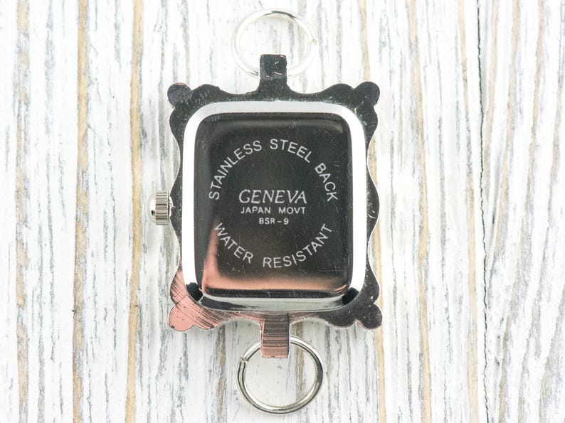 K&ouml;nnte beinhalten: Eine silberfarbene Geneva-Uhr mit rechteckigem Zifferblatt und Edelstahlr&uuml;ckseite. Die Uhr hat einen dekorativen Rahmen und einen Schl&uuml;sselring-Aufsatz. Der Text auf dem Zifferblatt lautet "Geneva Japan Movt BSR-9 Water Resistant."