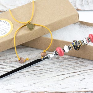 May include: A decorative letter opener with a silver blade and a beaded handle. The handle features pink, black, and clear beads, along with silver accents. A yellow cord with a heart charm and wooden beads is attached to the box.