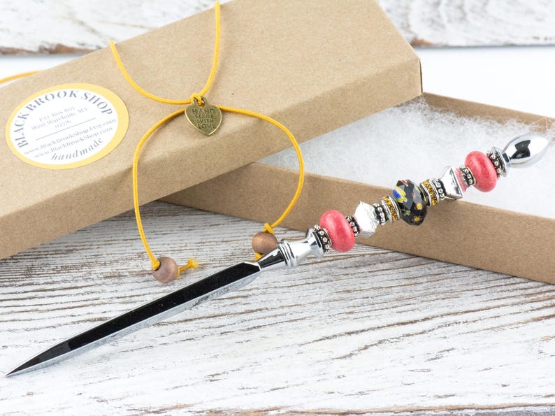 May include: A decorative letter opener with a silver blade and a beaded handle. The handle features pink, black, and clear beads, along with silver accents. A yellow cord with a heart charm and wooden beads is attached to the box.
