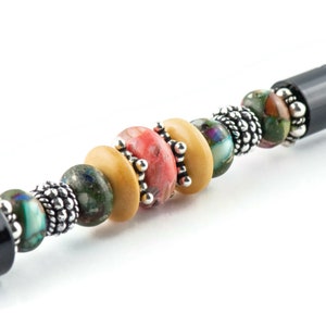 K&ouml;nnte beinhalten: Ein schwarzer Stift mit einem dekorativen Perlenmuster. Die Perlen sind aus Holz, Stein und Metall. Die Perlen sind in den Farben Braun, Gr&uuml;n, Rosa und Blau.