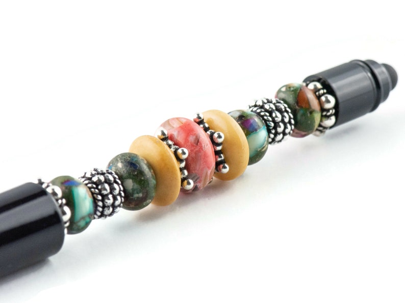 K&ouml;nnte beinhalten: Ein schwarzer Stift mit einem dekorativen Perlenmuster. Die Perlen sind aus Holz, Stein und Metall. Die Perlen sind in den Farben Braun, Gr&uuml;n, Rosa und Blau.