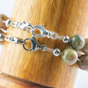 Pu&ograve; includere: Due braccialetti di perline con chiusure argentate. Le perline sono un mix di colori verdi, marroni e beige. I braccialetti sono esposti su un supporto di legno su sfondo bianco.