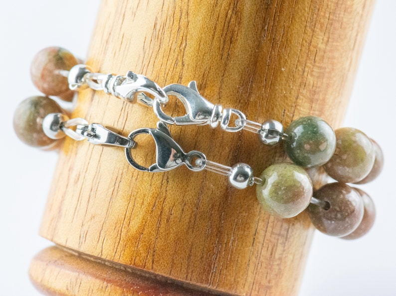 Pu&ograve; includere: Due braccialetti di perline con chiusure argentate. Le perline sono un mix di colori verdi, marroni e beige. I braccialetti sono esposti su un supporto di legno su sfondo bianco.