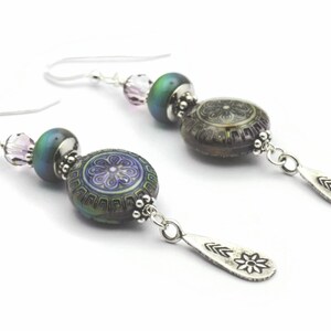 May include: Silver dangle earrings featuring a purple and green iridescent glass bead, a smaller pink crystal bead, and a silver charm with a flower design.