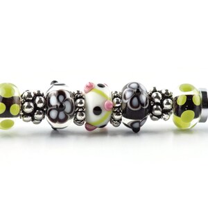 K&ouml;nnte beinhalten: Ein silbernes Charm-Armband mit einer Reihe dekorativer Perlen. Die Perlen weisen verschiedene Designs auf, darunter schwarz-wei&szlig;e Blumenmuster, gr&uuml;ne Tupfen und eine wei&szlig;e Perle mit rosa Akzenten. Zwischen den Perlen befinden sich silberne Abstandshalter.