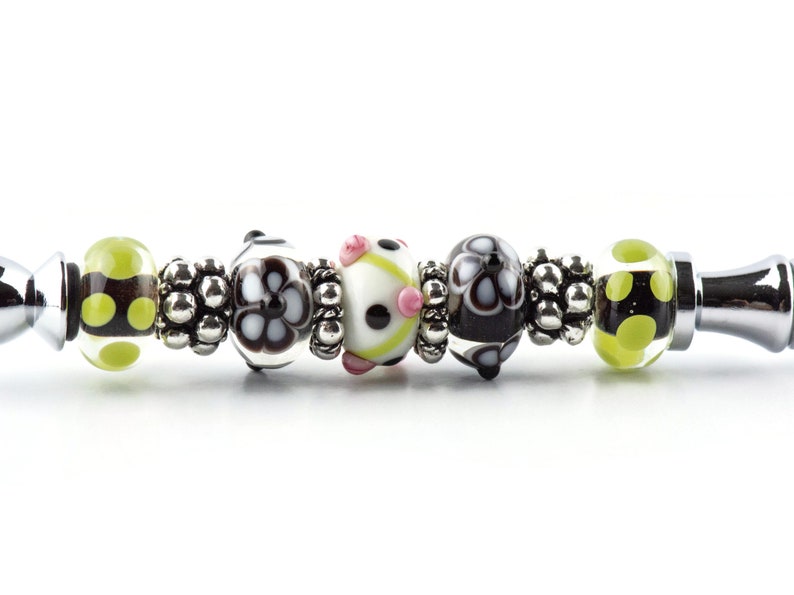 K&ouml;nnte beinhalten: Ein silbernes Charm-Armband mit einer Reihe dekorativer Perlen. Die Perlen weisen verschiedene Designs auf, darunter schwarz-wei&szlig;e Blumenmuster, gr&uuml;ne Tupfen und eine wei&szlig;e Perle mit rosa Akzenten. Zwischen den Perlen befinden sich silberne Abstandshalter.