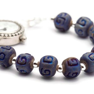 Pu&ograve; includere: Un orologio da tasca d'argento con quadrante bianco e lancette nere &egrave; attaccato a un braccialetto fatto di perline blu scuro con un motivo a spirale.