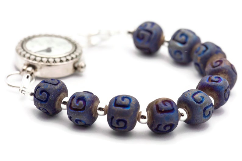 Pu&ograve; includere: Un orologio da tasca d'argento con quadrante bianco e lancette nere &egrave; attaccato a un braccialetto fatto di perline blu scuro con un motivo a spirale.