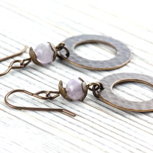 Puede incluir: Un par de pendientes hechos a mano con una cuenta de piedra preciosa de color p&uacute;rpura claro y un c&iacute;rculo de metal martillado. Los pendientes tienen un gancho de color bronce y est&aacute;n sobre un fondo de madera blanca.