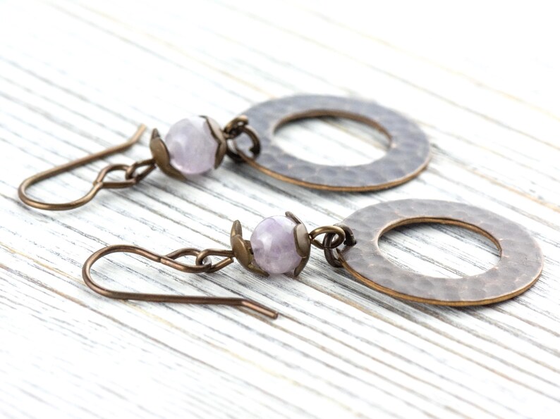 Puede incluir: Un par de pendientes hechos a mano con una cuenta de piedra preciosa de color p&uacute;rpura claro y un c&iacute;rculo de metal martillado. Los pendientes tienen un gancho de color bronce y est&aacute;n sobre un fondo de madera blanca.