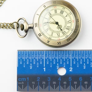 Op de afbeelding: Antieke messing zakhorloge met Romeinse cijfers op de wijzerplaat, bevestigd aan een ketting. Een blauwe liniaal met centimeter- en millimeter markeringen is eronder geplaatst. De wijzerplaat heeft een diameter van ongeveer 4,4 cm.