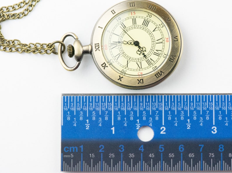 Op de afbeelding: Antieke messing zakhorloge met Romeinse cijfers op de wijzerplaat, bevestigd aan een ketting. Een blauwe liniaal met centimeter- en millimeter markeringen is eronder geplaatst. De wijzerplaat heeft een diameter van ongeveer 4,4 cm.