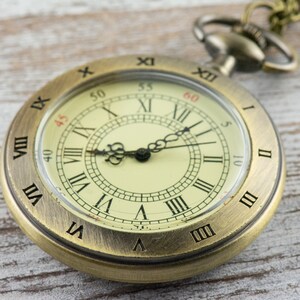 Op de afbeelding: Een vintage zakhorloge met een messingkleurige kast en een cr&egrave;mekleurige wijzerplaat. Het horloge heeft zwarte Romeinse cijfers, zwarte wijzers en een kleine secondenwijzer. Het horloge is bevestigd aan een ketting.