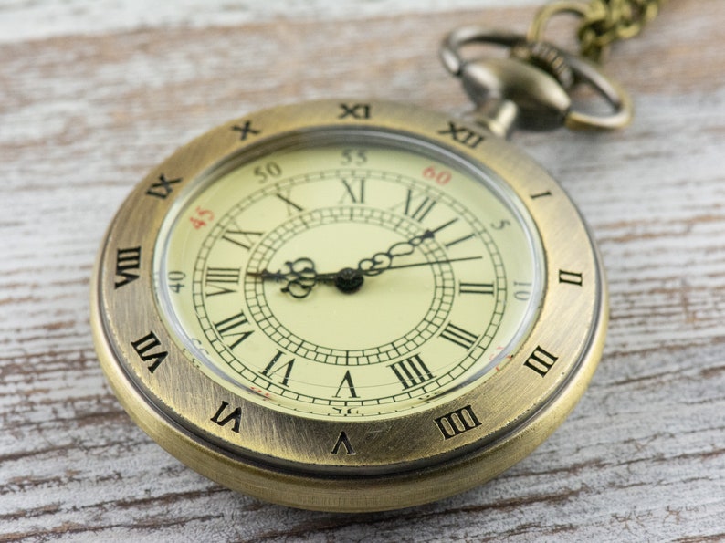Op de afbeelding: Een vintage zakhorloge met een messingkleurige kast en een cr&egrave;mekleurige wijzerplaat. Het horloge heeft zwarte Romeinse cijfers, zwarte wijzers en een kleine secondenwijzer. Het horloge is bevestigd aan een ketting.