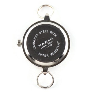 May include: A round silver-tone watch case with a black back featuring the text STAINLESS STEEL BACK, NARMI, JAPAN MOVT, L-133, and WATER RESISTANT. The watch includes a side crown and two silver metal rings attached to the top and bottom lugs.