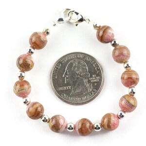 May include: A pink and brown patterned gemstone bracelet with silver beads and a silver clasp. The bracelet is about the size of a US quarter.