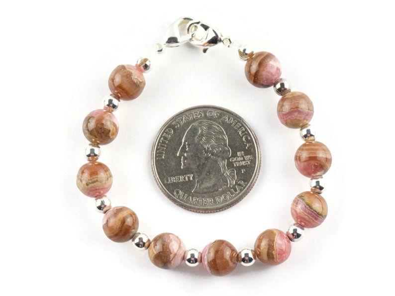 May include: A pink and brown patterned gemstone bracelet with silver beads and a silver clasp. The bracelet is about the size of a US quarter.