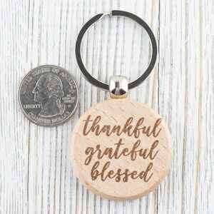 May include: A wooden keychain with a round, light brown tag that says "thankful grateful blessed" in a cursive font. The keychain is attached to a silver metal ring.