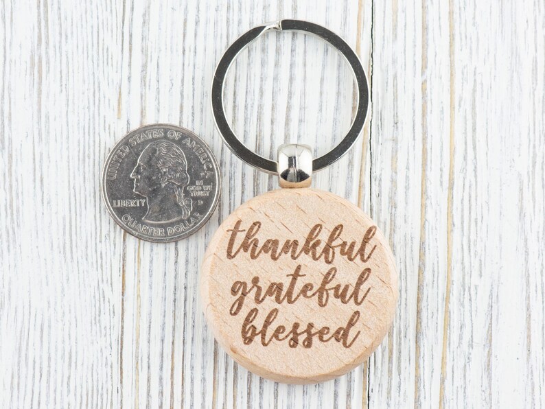 May include: A wooden keychain with a round, light brown tag that says "thankful grateful blessed" in a cursive font. The keychain is attached to a silver metal ring.