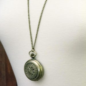 K&ouml;nnte beinhalten: Antike, messingfarbene Taschenuhr-Anh&auml;nger-Halskette. Der runde Uhrenanh&auml;nger hat ein verziertes Blumenmuster. Die Halskette hat eine passende Messingfarbe. Die Uhr wird auf einer wei&szlig;en Oberfl&auml;che pr&auml;sentiert.