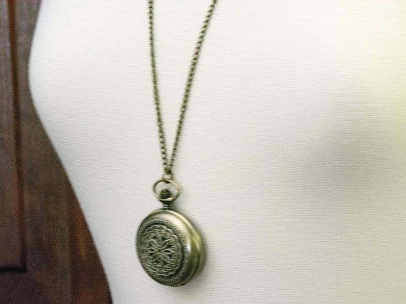 K&ouml;nnte beinhalten: Antike, messingfarbene Taschenuhr-Anh&auml;nger-Halskette. Der runde Uhrenanh&auml;nger hat ein verziertes Blumenmuster. Die Halskette hat eine passende Messingfarbe. Die Uhr wird auf einer wei&szlig;en Oberfl&auml;che pr&auml;sentiert.