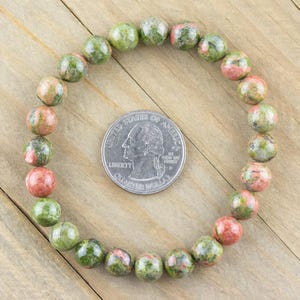 May include: A green, pink, and brown gemstone bracelet with round beads. The bracelet is laid out on a wooden surface next to a US quarter.
