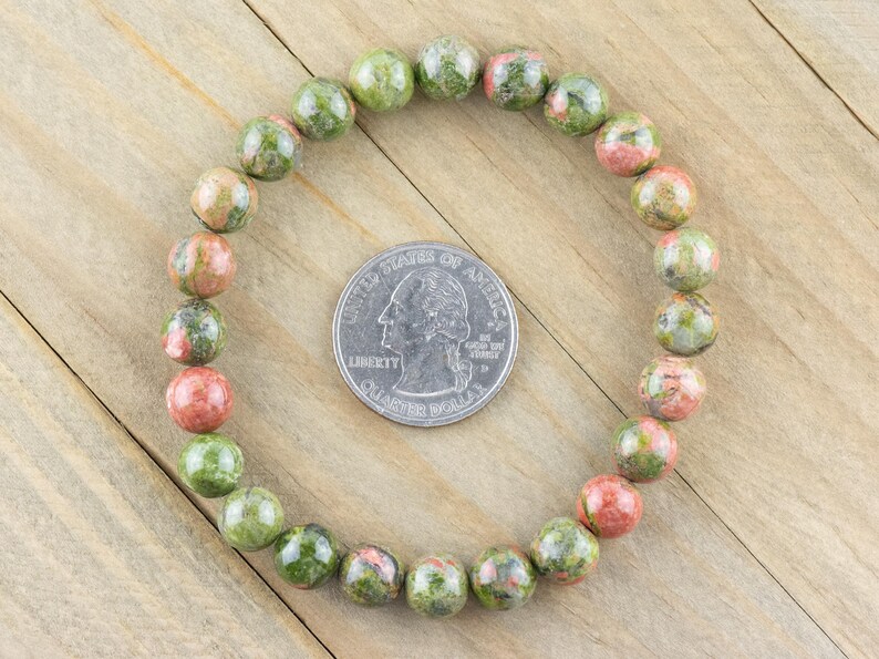 May include: A green, pink, and brown gemstone bracelet with round beads. The bracelet is laid out on a wooden surface next to a US quarter.