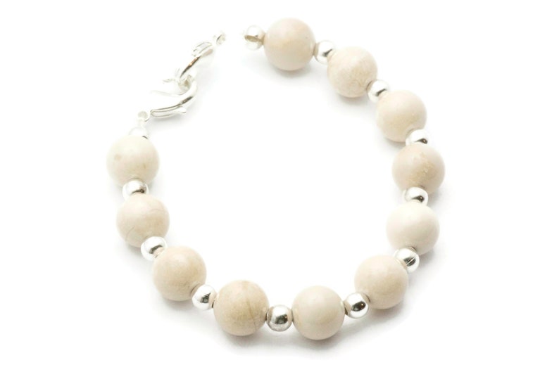 Op de afbeelding: Een witte kralen armband met zilveren accenten. De armband is gemaakt van ronde, gladde kralen en heeft een zilveren sluiting.