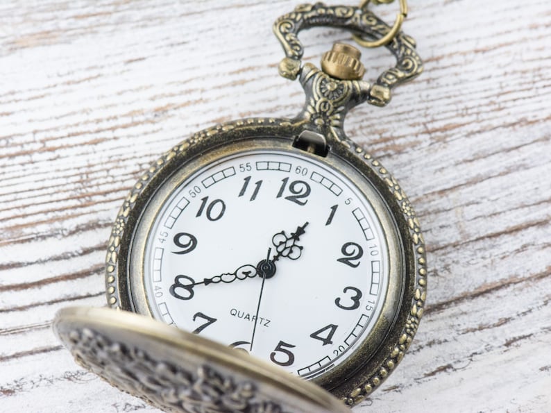 May include: A vintage-style pocket watch with a gold-toned metal case and a white face. The watch has black hands and black Roman numeral hour markers. The watch face also has a small black second hand and the word "QUARTZ" printed in black.