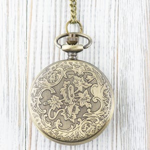 May include: A gold pocket watch with a floral design on the front. The watch is hanging from a chain.