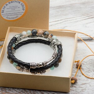 May include: A set of three bracelets with black leather cord, silver beads, and gemstone beads. The bracelets are in a brown box with a yellow ribbon. A necklace with a silver heart pendant that says "Made for You" is also included.