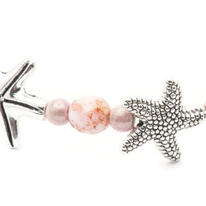 K&ouml;nnte beinhalten: Ein Armband mit silbernen Seestern-Anh&auml;ngern und rosa- und mauvefarbenen Perlen. Das Armband hat ein maritimes Thema, wobei die Seesterne ein dekoratives Element hinzuf&uuml;gen. Die Perlen haben ein gesprenkeltes Aussehen.
