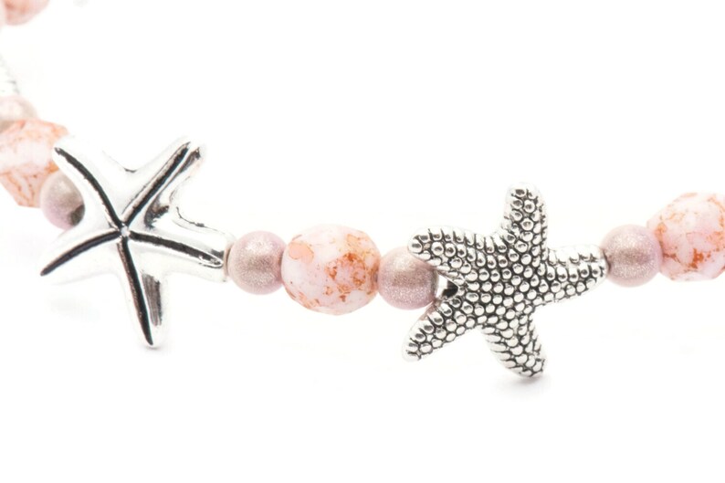 K&ouml;nnte beinhalten: Ein Armband mit silbernen Seestern-Anh&auml;ngern und rosa- und mauvefarbenen Perlen. Das Armband hat ein maritimes Thema, wobei die Seesterne ein dekoratives Element hinzuf&uuml;gen. Die Perlen haben ein gesprenkeltes Aussehen.