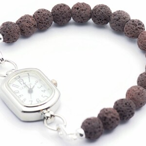 Puede incluir: Un reloj de pulsera con una esfera rectangular plateada y una esfera blanca. El reloj est&aacute; conectado a una pulsera hecha de cuentas de roca volc&aacute;nica marr&oacute;n oscuro y porosa. La correa del reloj se asegura con un cierre plateado.