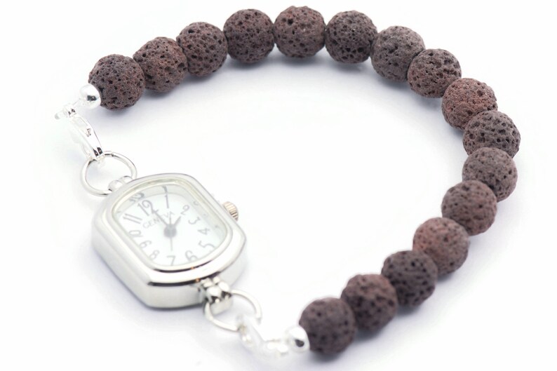 Puede incluir: Un reloj de pulsera con una esfera rectangular plateada y una esfera blanca. El reloj est&aacute; conectado a una pulsera hecha de cuentas de roca volc&aacute;nica marr&oacute;n oscuro y porosa. La correa del reloj se asegura con un cierre plateado.