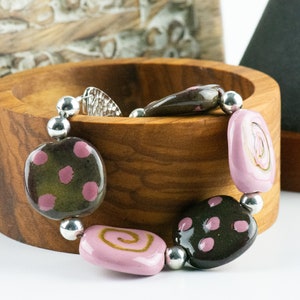 May include: A bracelet made of brown and pink ceramic beads with silver accents. The beads are patterned with polka dots and swirls.